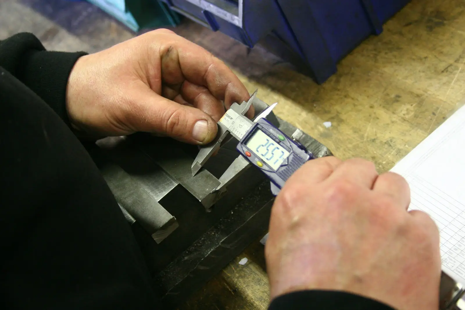 A worker measuring a machined steel part with a digital caliper at a bench