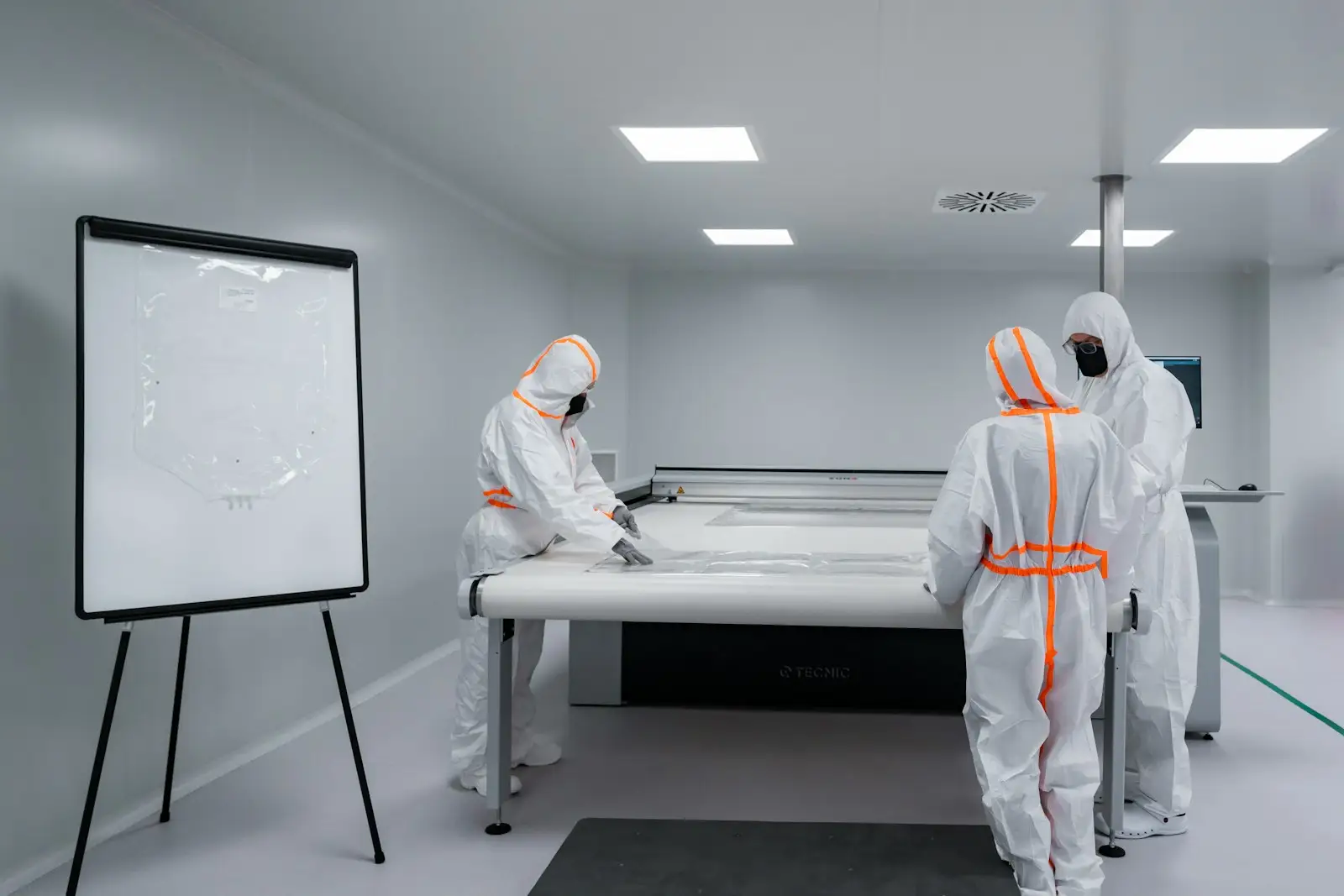 Three gowned operators at a cleanroom work table with a reference diagram on an easel