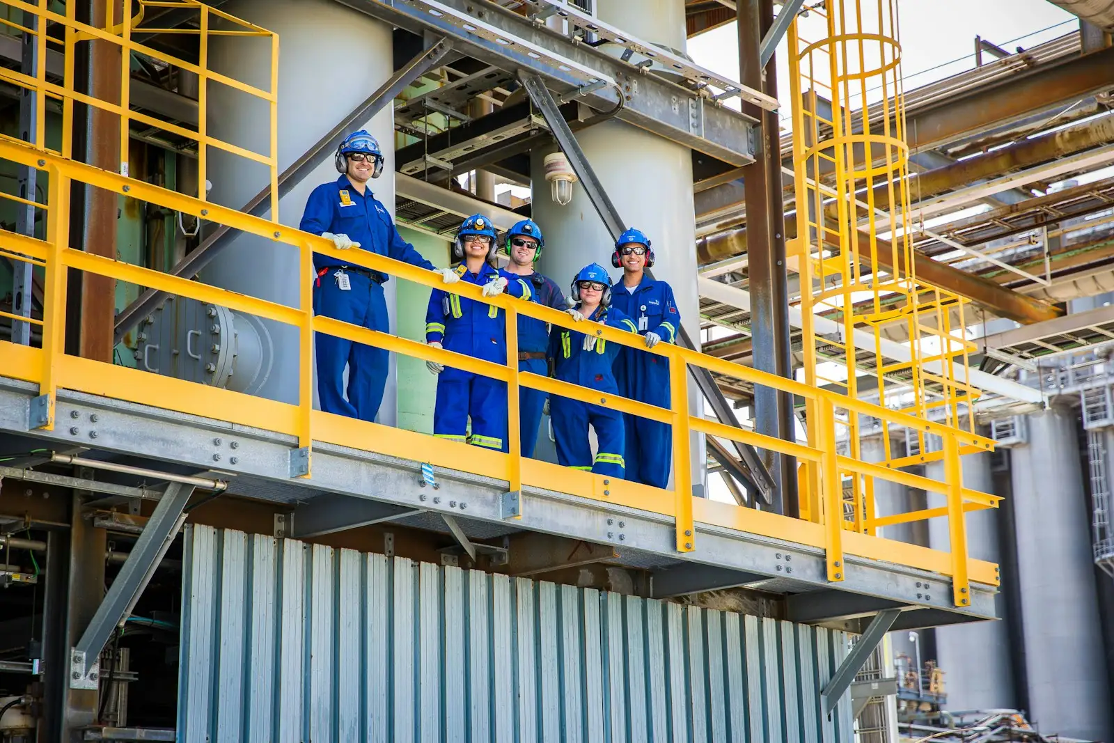 Five plant workers in coveralls and hard hats on an elevated platform