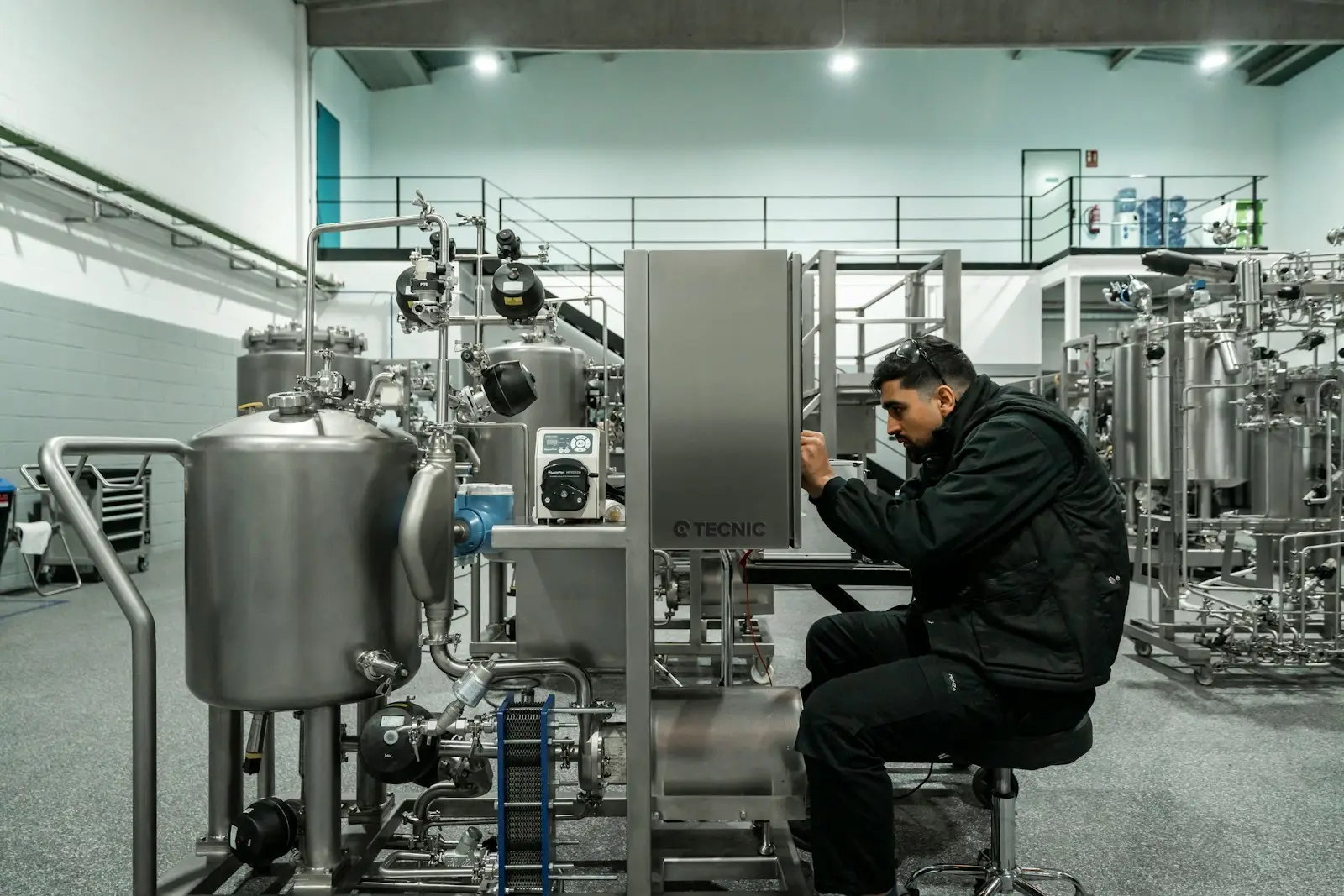 A technician servicing an open stainless steel equipment cabinet among tanks and piping