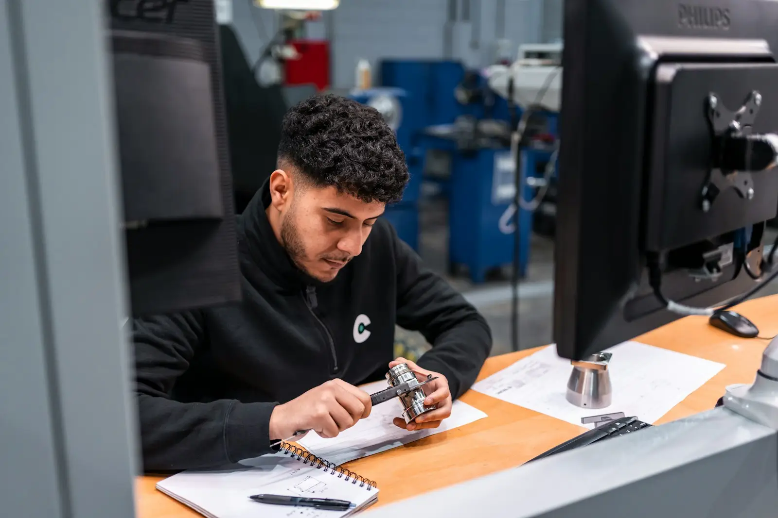 A technician checking a metal part with calipers, notebook and sketches beside him