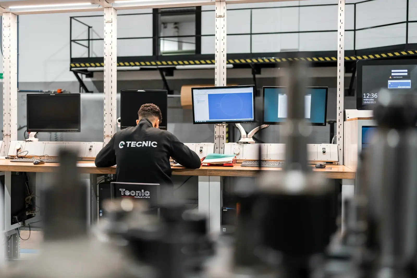 A technician at a mezzanine workstation reviewing drawings on several monitors above the shop floor