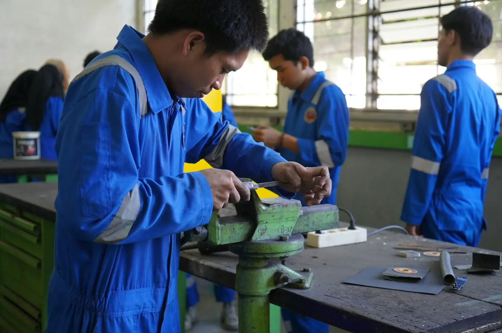 A trainee in blue coveralls filing metal at a bench vice, others working behind