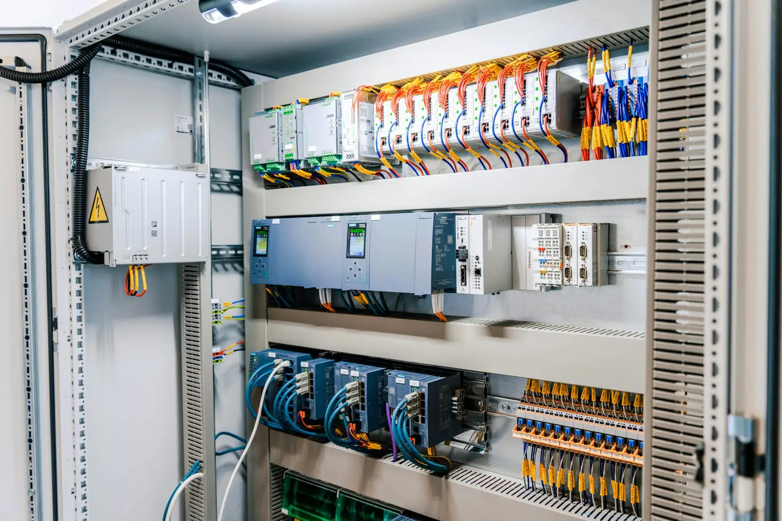 An open control cabinet with PLC modules and DIN rail I/O on color coded wiring