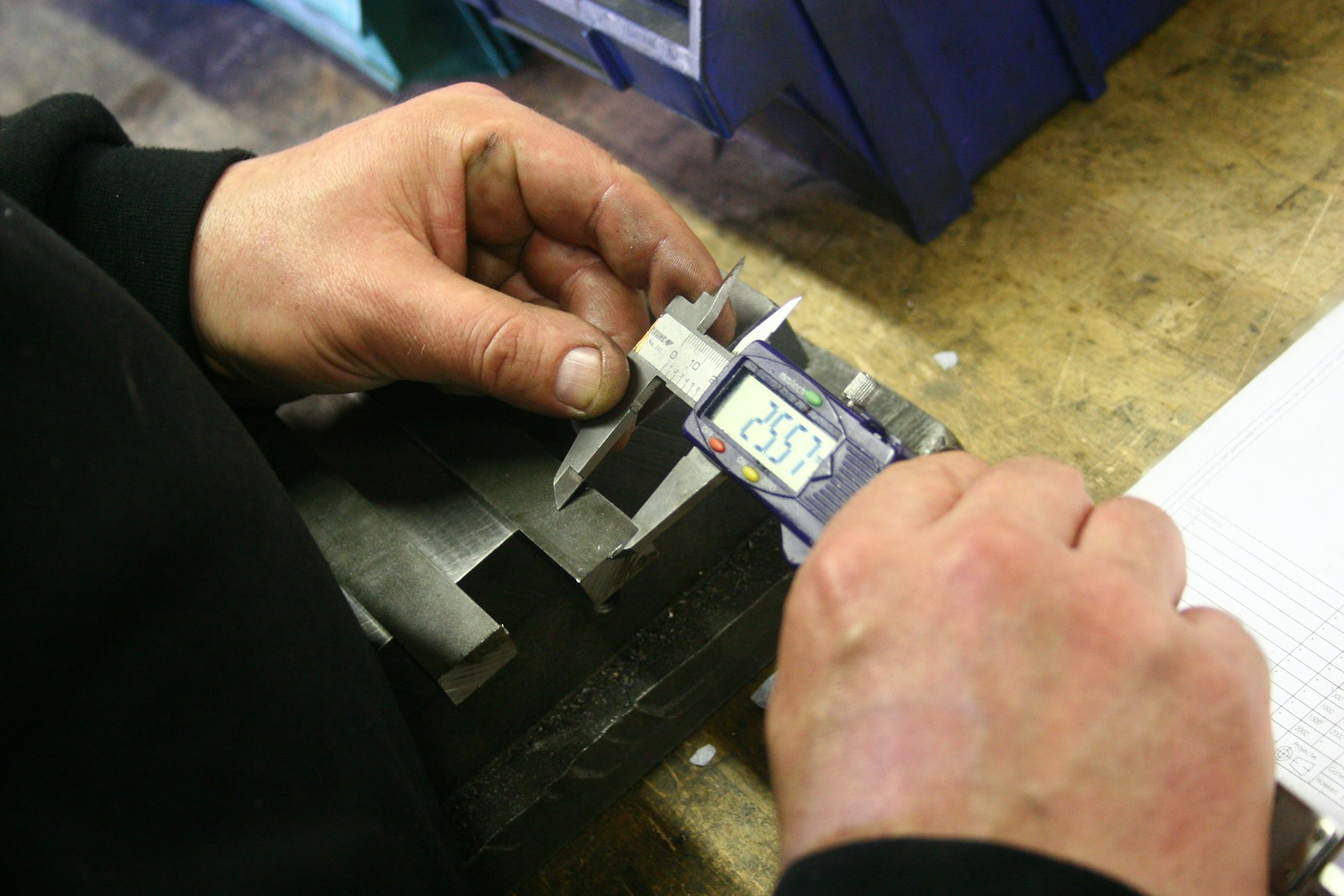 A worker measuring a machined steel part with a digital caliper at a bench